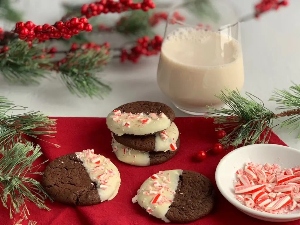 4 chocolate cookies dipped in melted white chocolate with crushed candy canes on them next to a glass of almond milk. 