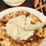 A white plate with 2 large waffles on it. The waffles have a cinnamon swirl mixture in the them and topped with a creamy frosting.