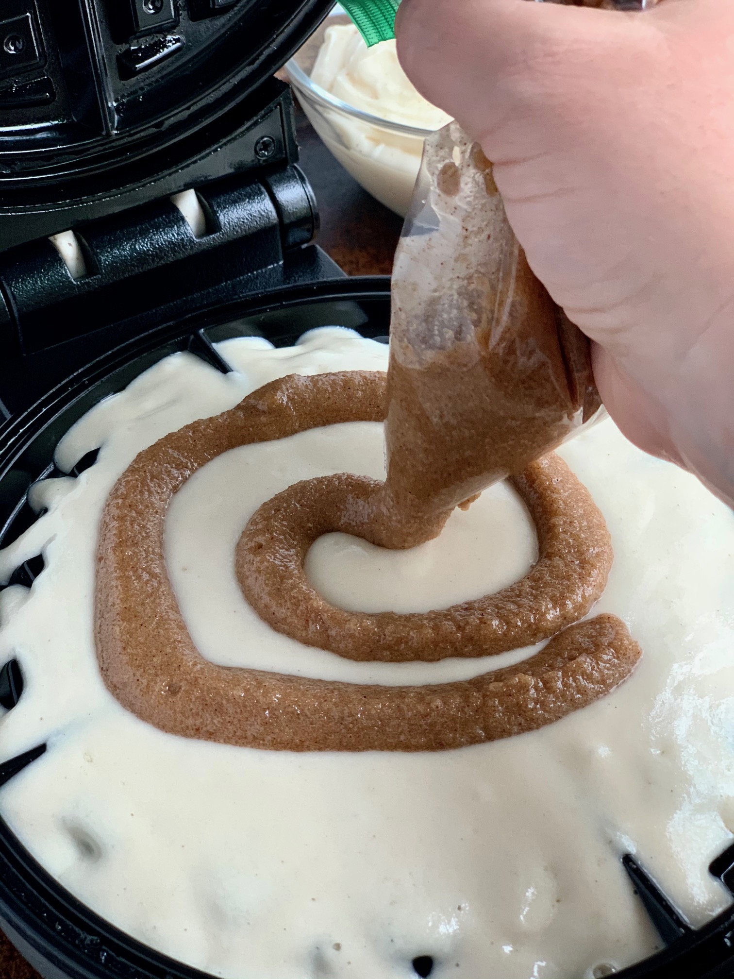 A waffle maker with waffle mix on it. A hand is piping a cinnamon swirl mixture from a zip lock bag onto the waffle mix.