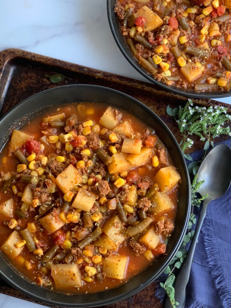 A large black bowl filled with tender potatoes, chunks of ground beef, corn, green beans, fire roasted tomato, broth, and a tomato juice combination.