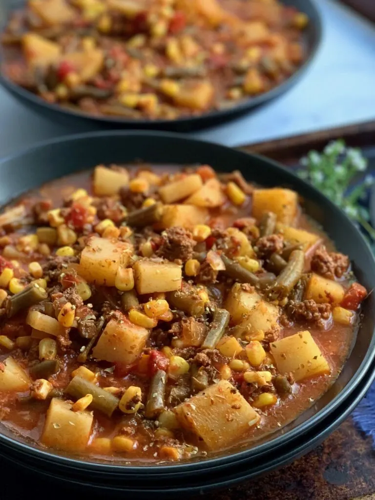 A large black bowl filled with tender potatoes, chunks of ground beef, corn, green beans, fire roasted tomato, broth, and a tomato juice combination.
