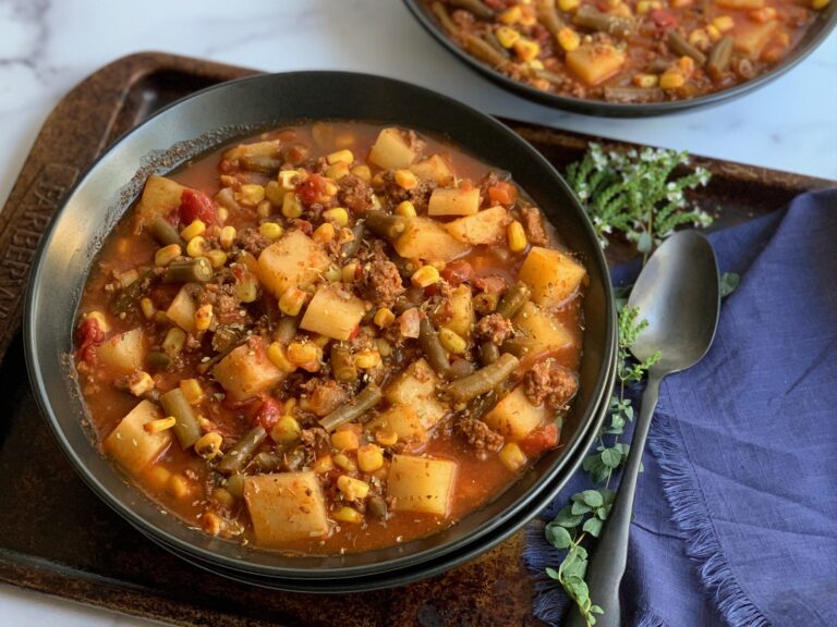 A large black bowl filled with tender potatoes, chunks of ground beef, corn, green beans, fire roasted tomato, broth, and a tomato juice combination.