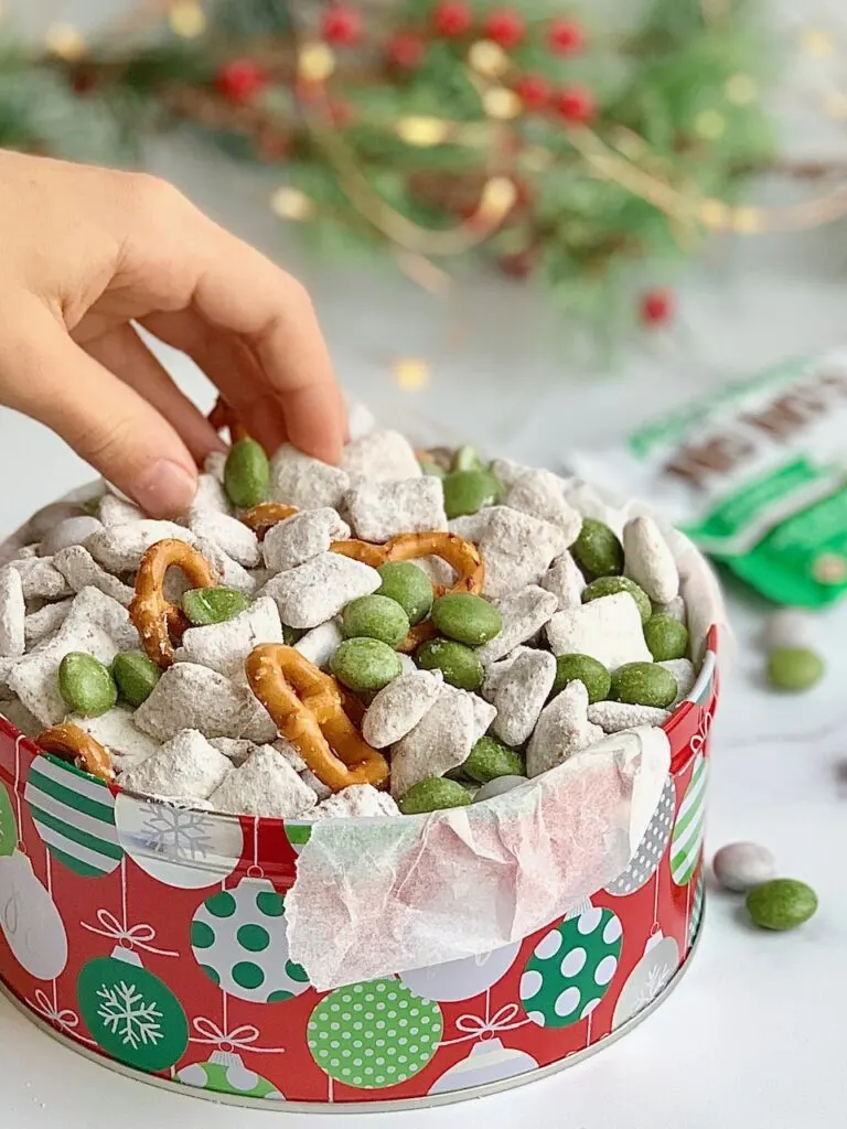 A holiday tin filled with homemade puppy chow, pretzels twists, and No Whey Peppermint No No's and a hand reaching in to grab some.