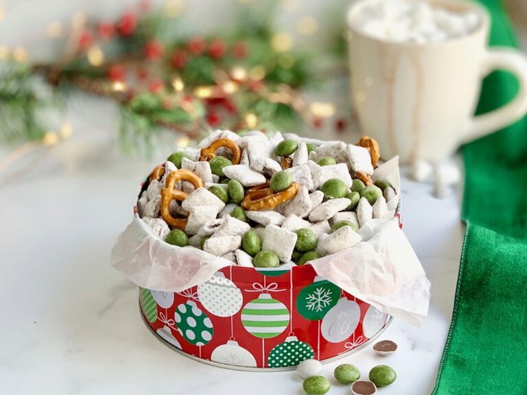 A large Christmas tin full of puppy chow, pretzels, and No Whey Foods Peppermint No No's