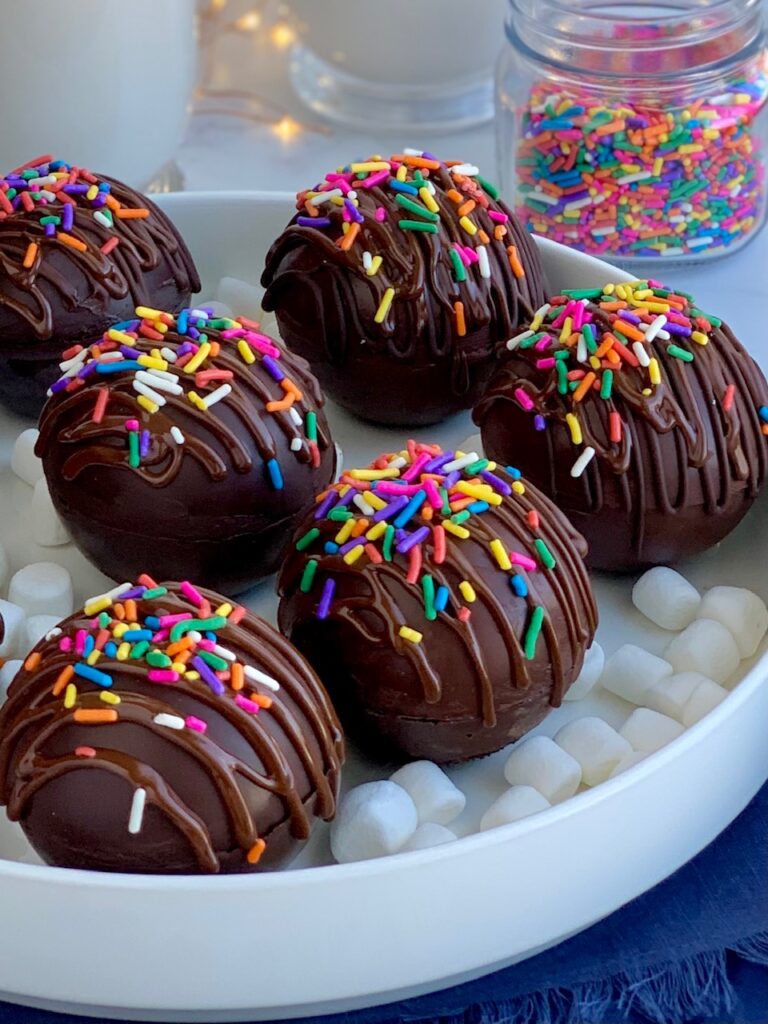 A platter full of thin chocolate balls stuffed with hot chocolate mix and mini marshmallows. The top of the balls are decorated with melted chocolate zig zagging across the top and topped with colorful sprinkles.