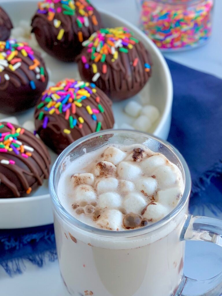 A platter full of thin chocolate balls stuffed with hot chocolate mix and mini marshmallows. The top of the balls are decorated with melted chocolate zig zagging across the top and topped with colorful sprinkles. A a mug full of hot chocolate and mini marshmallows.