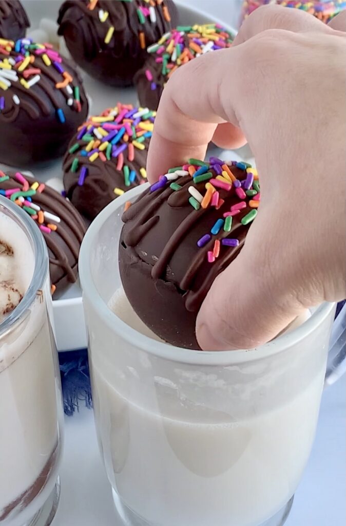 A thin chocolate ball stuffed with hot chocolate mix and mini marshmallows being placed into a mug of hot dairy free milk. The top of the ball is decorated with melted chocolate zig zagging across the top and topped with colorful sprinkles.