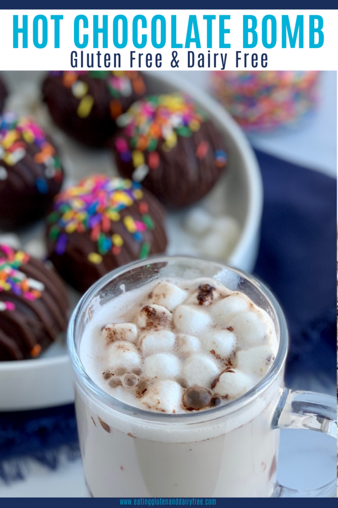 A platter full of thin chocolate balls stuffed with hot chocolate mix and mini marshmallows. The top of the balls are decorated with melted chocolate zig zagging across the top and topped with colorful sprinkles. A mug of warm hot chocolate topped with mini marshmallows.