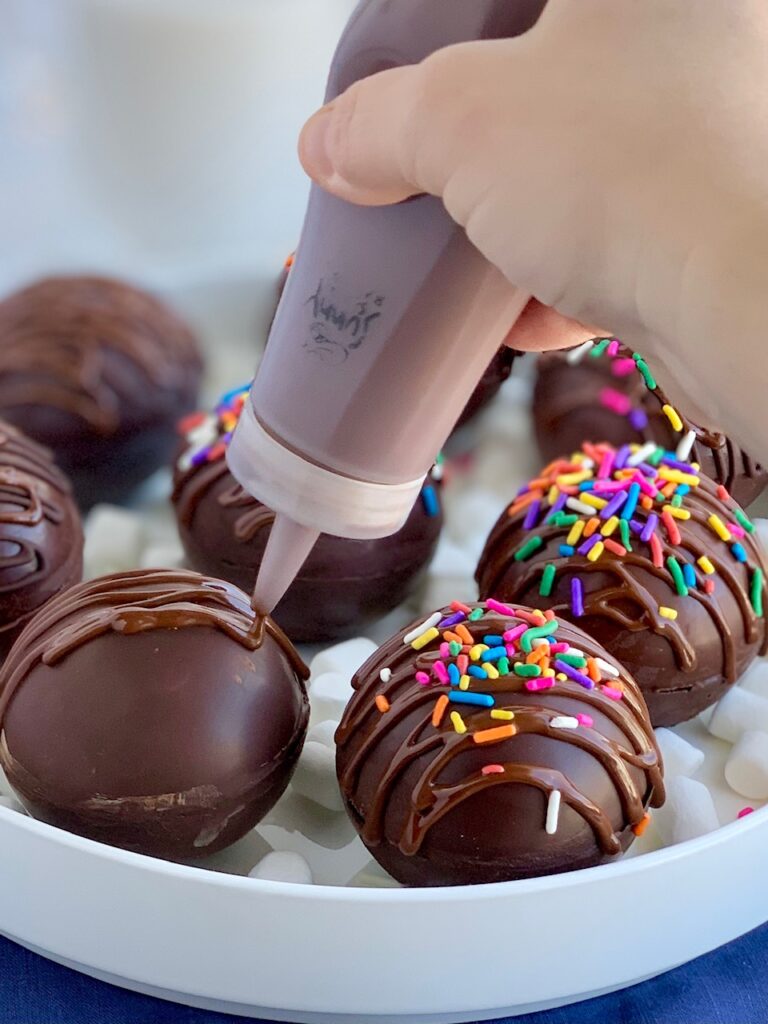 A platter full of thin chocolate balls stuffed with hot chocolate mix and mini marshmallows. The top of the balls are decorated with melted chocolate zig zagging across the top and topped with colorful sprinkles.