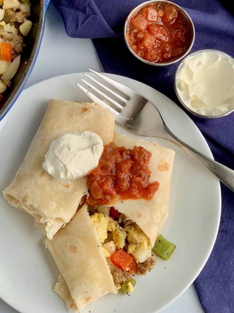 A white plate with 2 large breakfast burritos on it. One is cut open and you can see eggs, bell peppers, sausage, and hash browns. They are topped with dairy free sour cream and a dollop of salsa