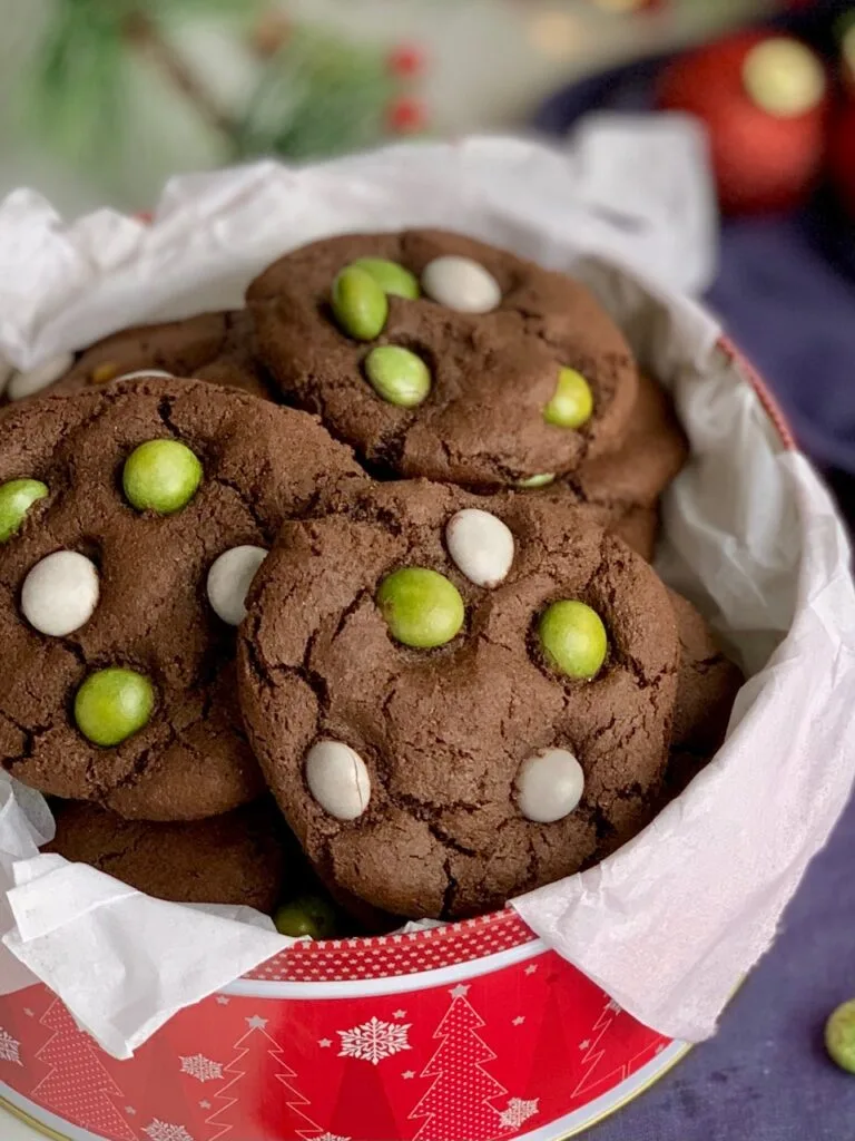 A festive red holiday tin filled with chocolate cookies featuring green and white candies on the top.