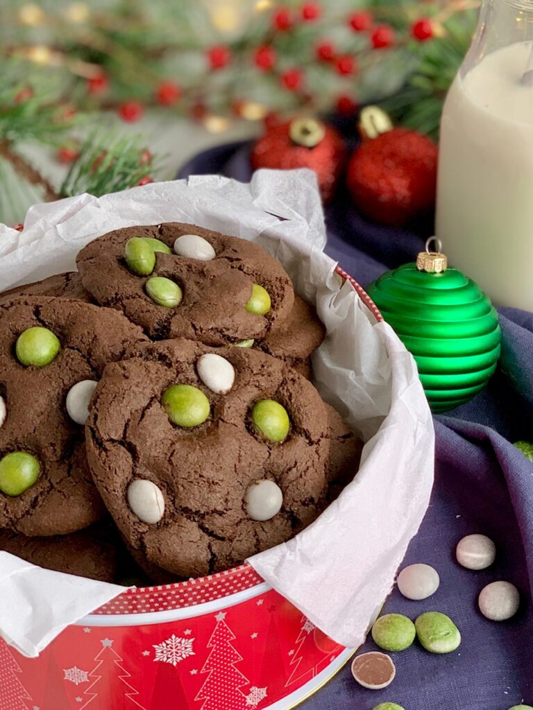 A festive red holiday tin filled with chocolate cookies featuring green and white candies on the top.