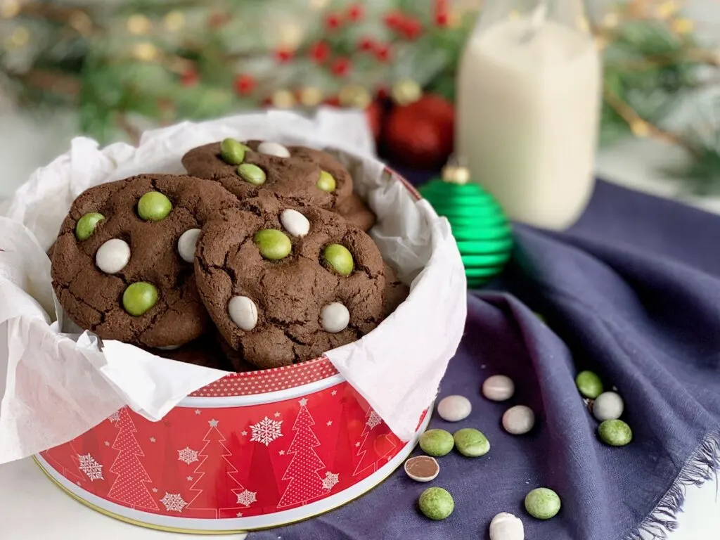 A festive red holiday tin filled with chocolate cookies featuring green and white candies on the top.