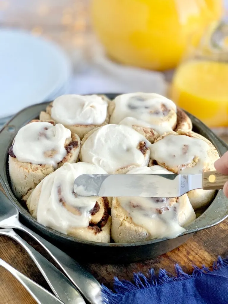 A pie plate filled with soft, gooey cinnamon rolls and topped with a decadent creamy dairy free cream cheese frosting next to serving plates and  glass of orange juice. An angled spatula is spreading around the cream chees frosting.