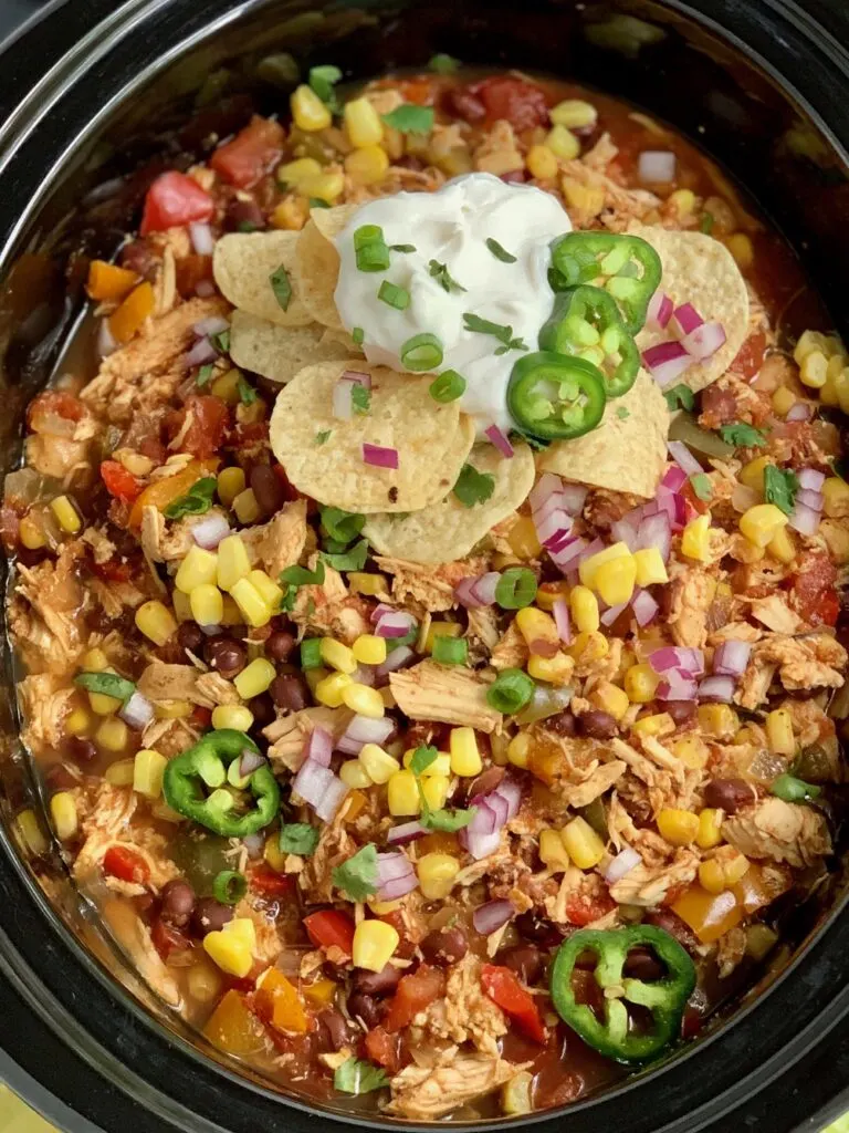 A crock-pot full of taco soup including shredded chicken, black beans, corn, onion, bell peppers, and lots of extra toppings.