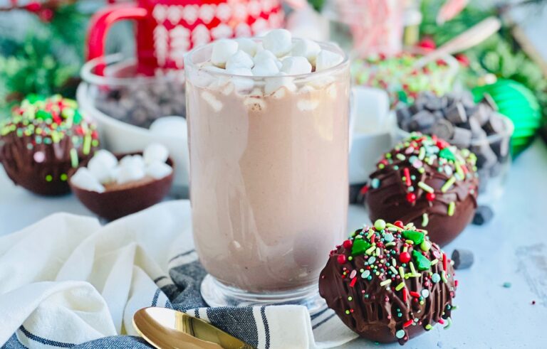A glass mug filled with steamy hot chocolate and mini white marshmallows on top. Next to a golden colored spoon and two hot chocolate bombs with chocolate drizzled across them and red, white, and green festive sprinkles on the top.