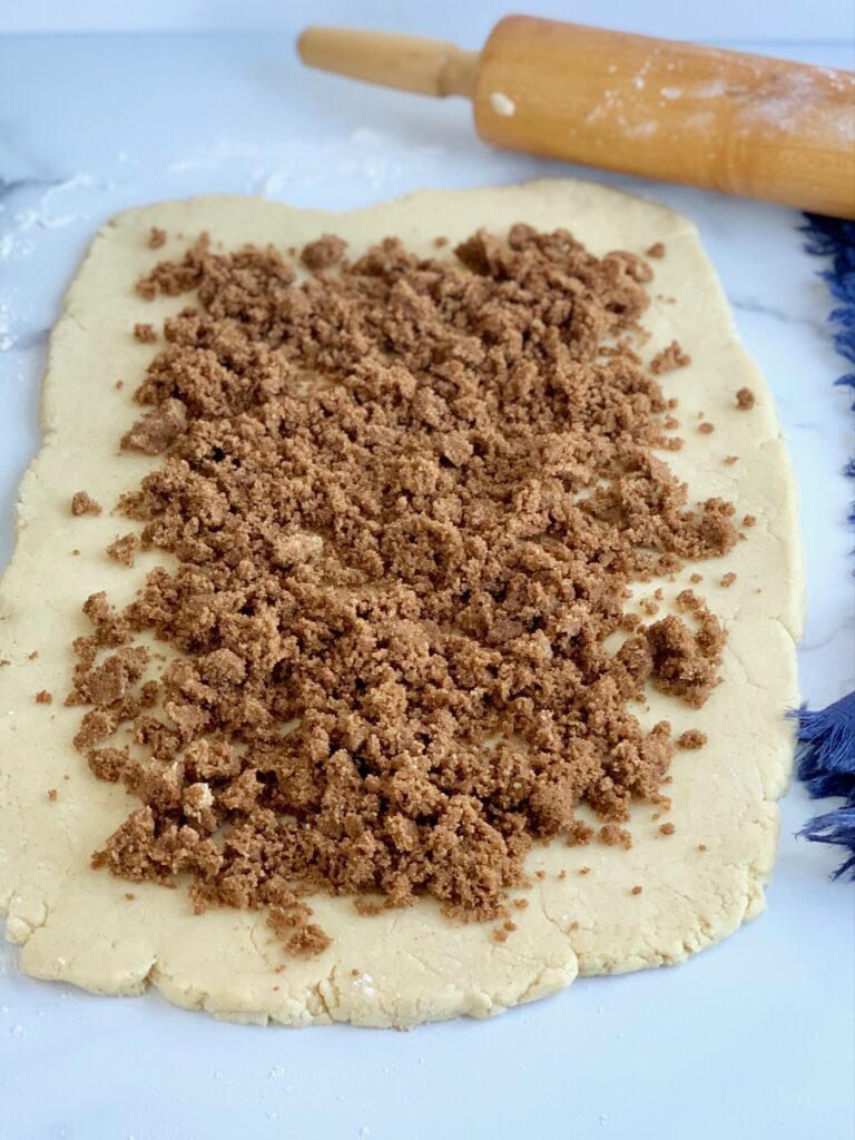 Rectangle shaped dough rolled out next to a rolling pin. It has a brown sugar, cinnamon, and melted butter mixture on top.