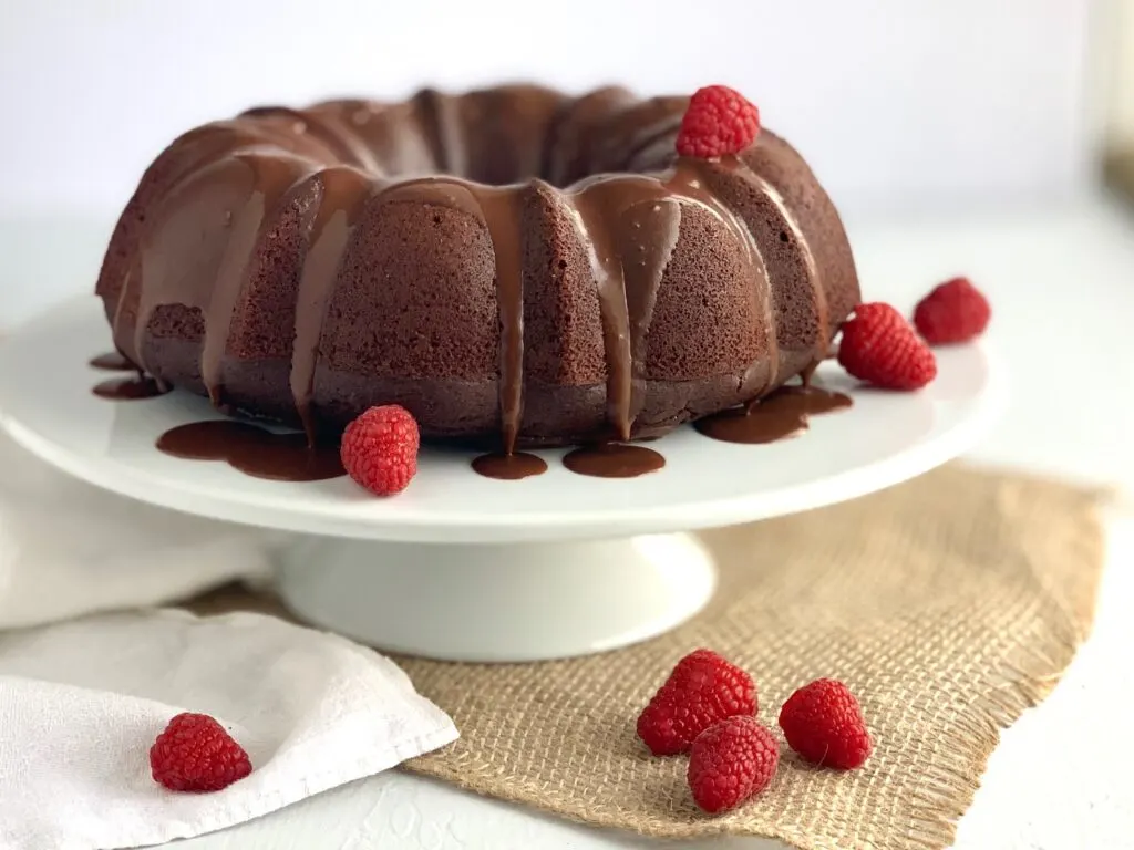 A large chocolate bundt cake on a white serving cake plate with a chocolate ganache running down the sides and random strawberries throughout. 