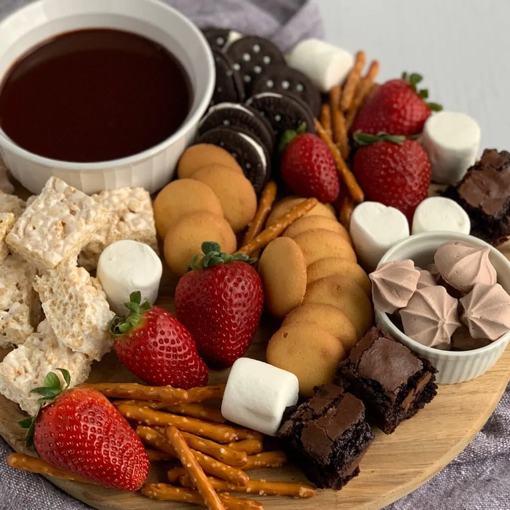 A serving board filled with amzing items such as mini rice krispies, mini brownie bites, safe oreos, pretzels, vanilla wafers, large marshmallows, strawberries and a small bowl of chocolate ganache dipping sauce. 