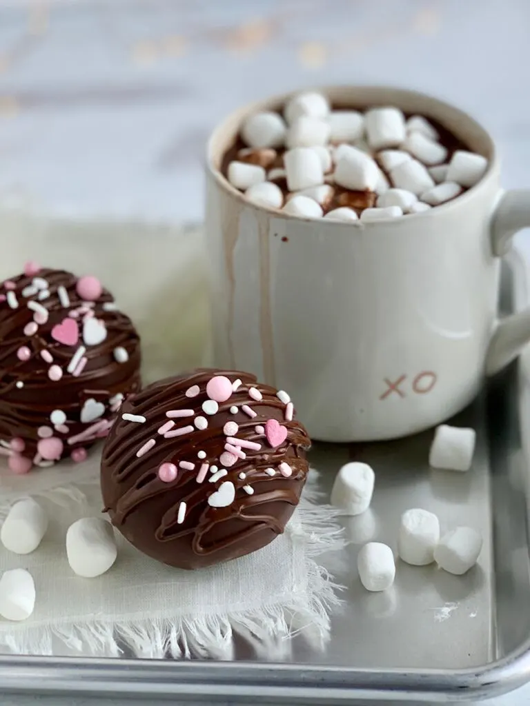 2 hollow chocolate balls filled with hot chocolate mix and sprinkles then decorated with drizzled chocolate on the outside and white and pink sprinkles. All next to a  mug of hot chocolate. 