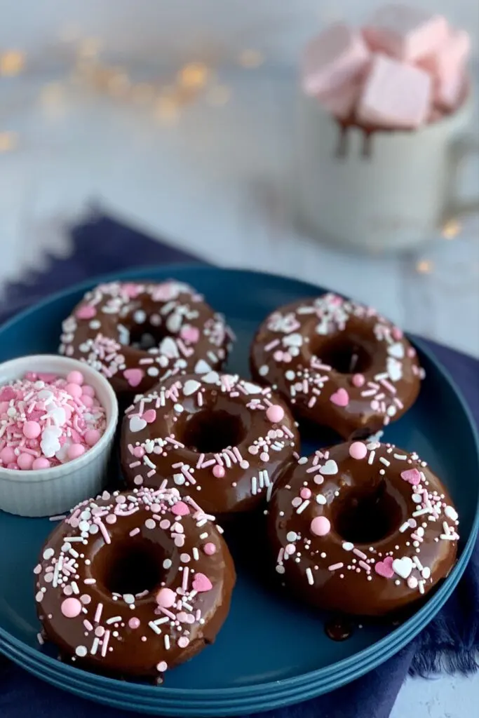 Chocolate Donuts with a chocolate ganache icing topped with white and pink sprinkles on a blue plate.