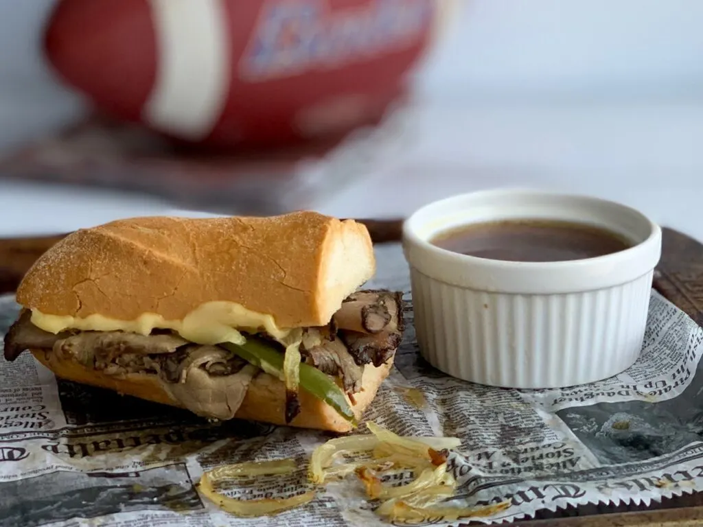 A toasted gluten free baguette with slices of roast beef, onions, and green peppers next to a bowl of au jus.