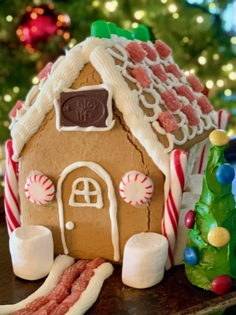 A gingerbread house decorated with frosting, candy, chocolate, marshmallows next to an upside down ice cream cone decorated as a tree with green frosting and colorful candies on it.