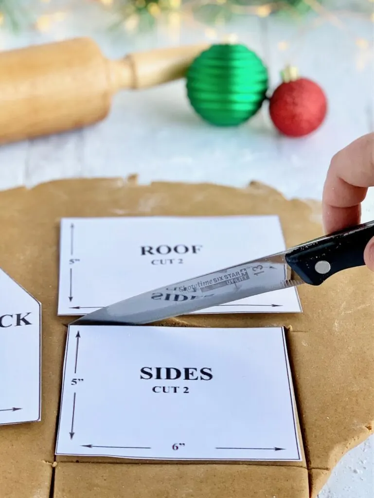 Gingerbread dough rolled out with 3 house panels laid on top-- the front and back panel, side panel, and roof panel. A knife is cutting out one of the side panels.