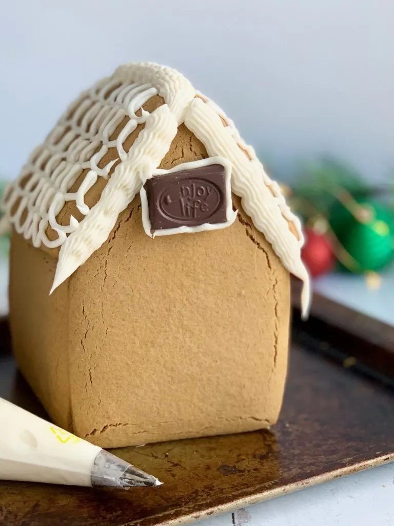 The gingerbread house is all glued together, and the roof has buttercream frosting on the very top and sides. The middle of the roof has decorative frosting scallops. An Enjoy Life candy bar piece is attached to the front of the house.