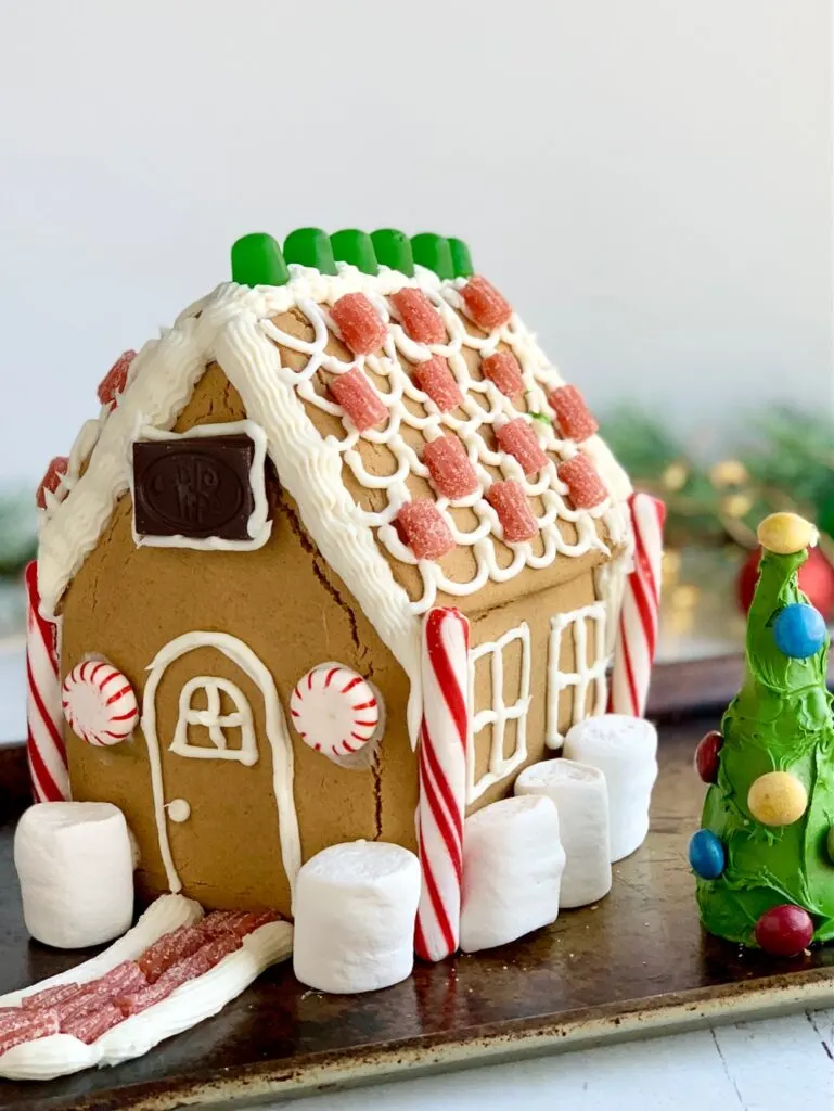 A gingerbread house decorated with frosting, candy, chocolate, marshmallows next to an upside down ice cream cone decorated as a tree with green frosting and colorful candies on it.