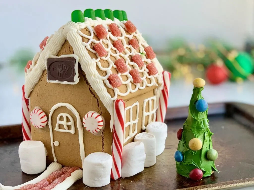 A gingerbread house decorated with frosting, candy, chocolate, marshmallows next to an upside down ice cream cone decorated as a tree with green frosting and colorful candies on it.