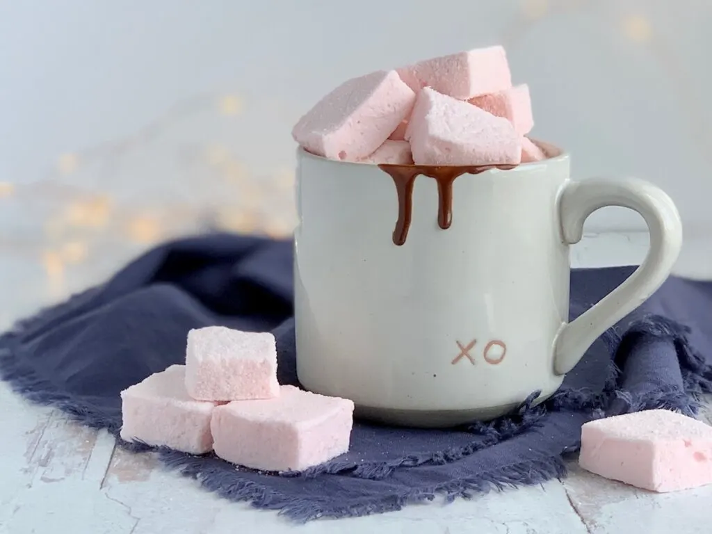 Soft fluffy pale pink square marshmallows on a table next to a mug of hot chocolate. 