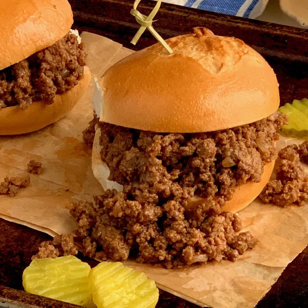 A massive helping of sloppy joes between a buttered bun with pickles on the side. 