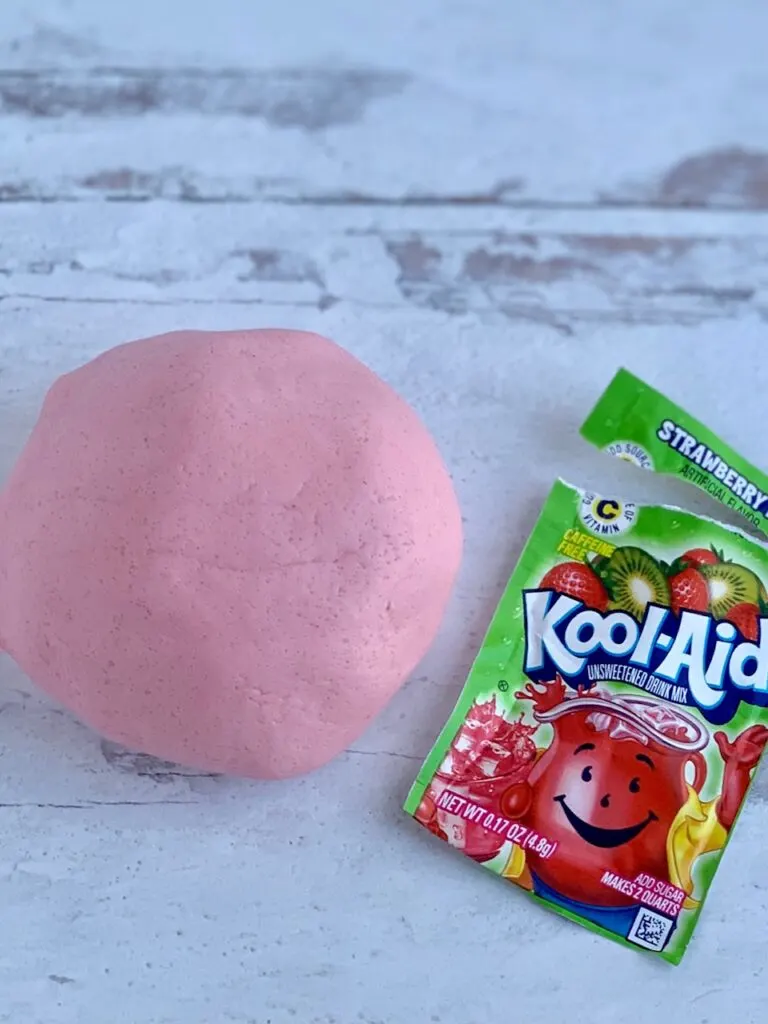 A round pink playdough ball on a countertop next to a packet of Kool-Aid.