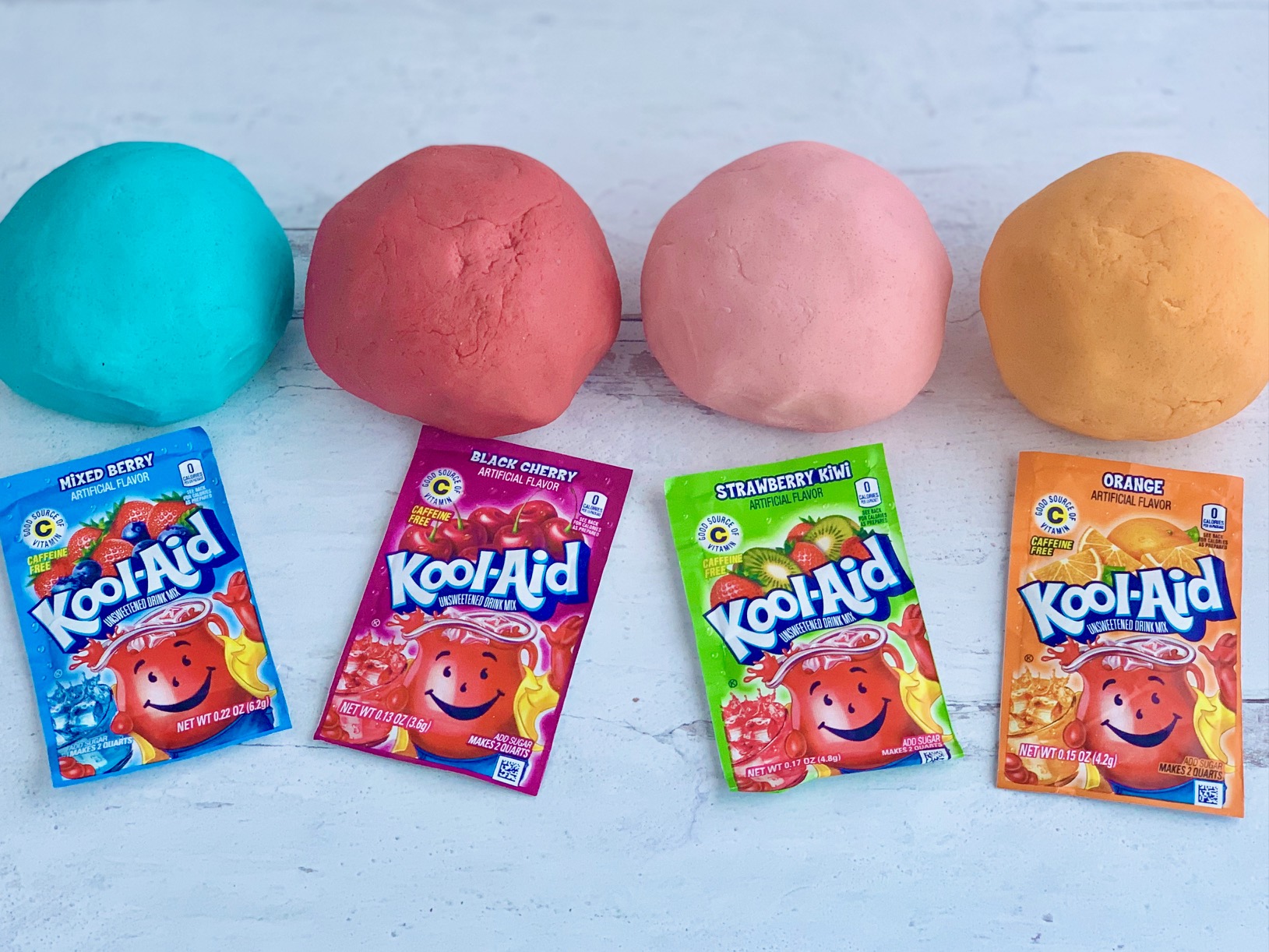 4 colorful balls of playdough next to matching packets of Kool-Aid.