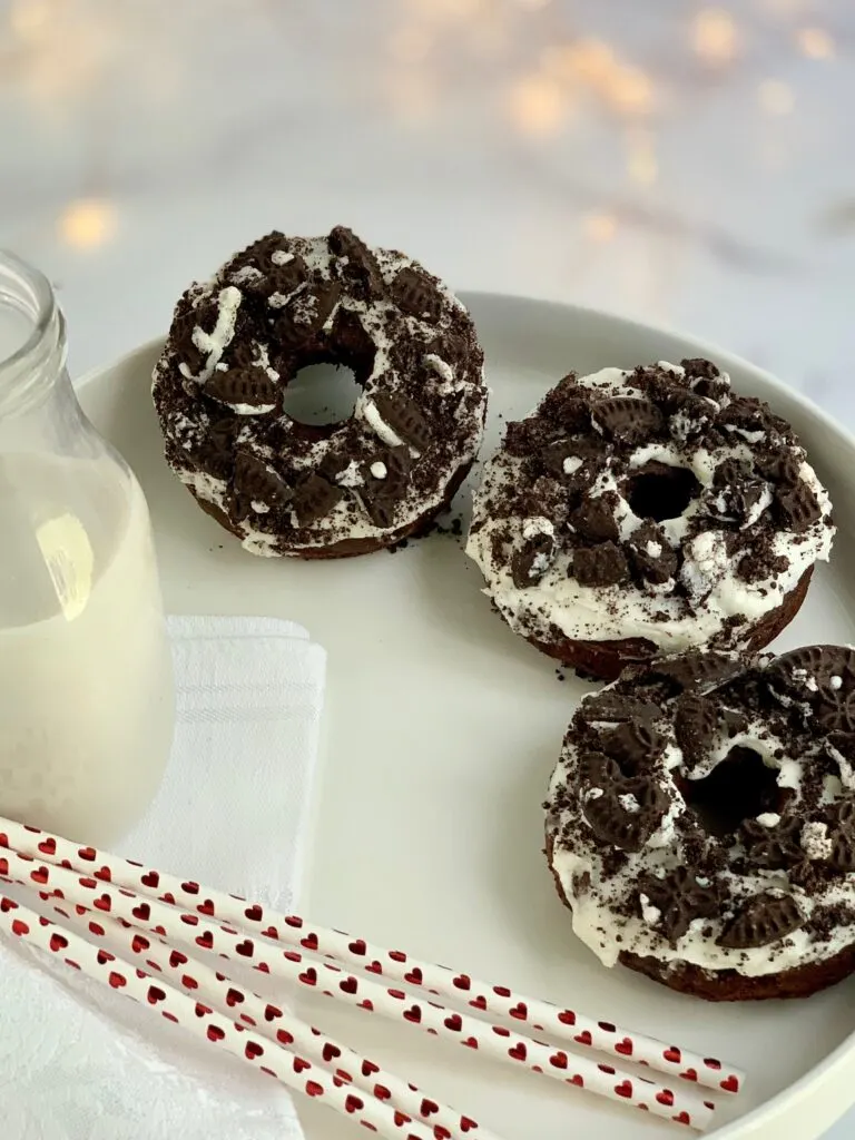 3 oreo donuts with a creamy white frosting and oreo crumbs sprinkled on top next to a glass of milk and 3 straws with red hearts on them.