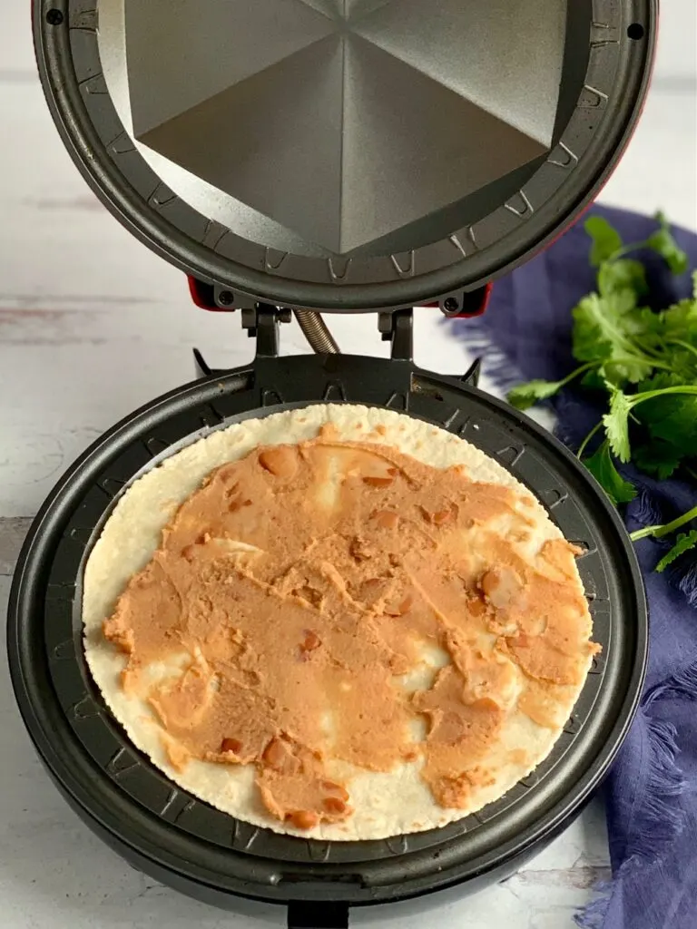 A quesadilla maker with a tortilla and refried beans spread on it.