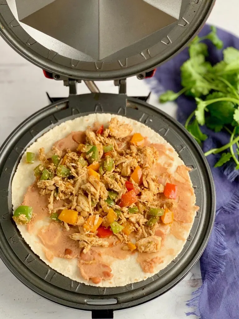 A quesadilla maker with a tortillas on it. On top of the tortillas is refried beans and the shredded chicken and bell pepper mixture. 