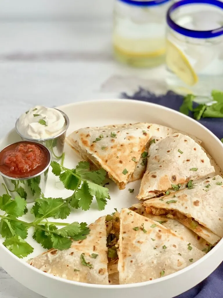 A white serving plate of triangle shaped quesadillas filled with chicken, bell peppers, and onions next to fresh salsa and dairy free sour cream.