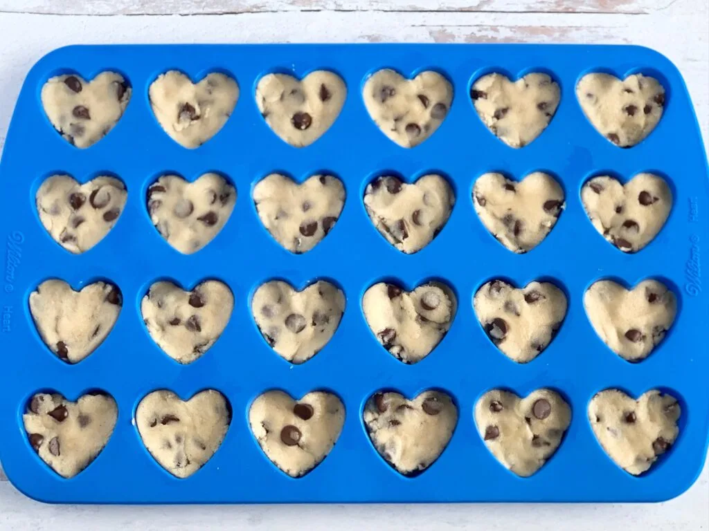 A blue silicone mold that has heart shaped cavities in it. Chocolate chip cookie dough is in the molds.