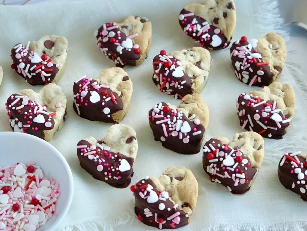 4 rows of mini heart shaped chocolate chip cookies that have been dipped halfway into melted chocolate then topped with pink, white, and red sprinkles.