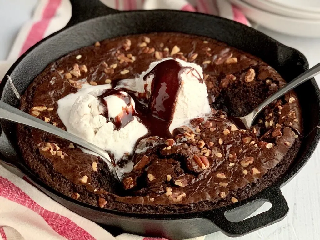 A cast iron skillet with baked brownies topped with nuts and 2 scoops of dairy free ice cream.