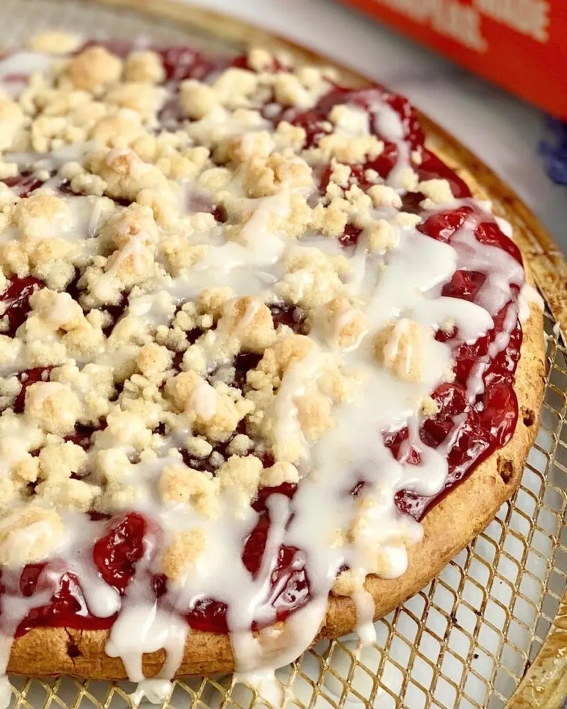 A pizza crust topped with cherry pie filling, a sugar crumble, and powdered sugar drizzle. 