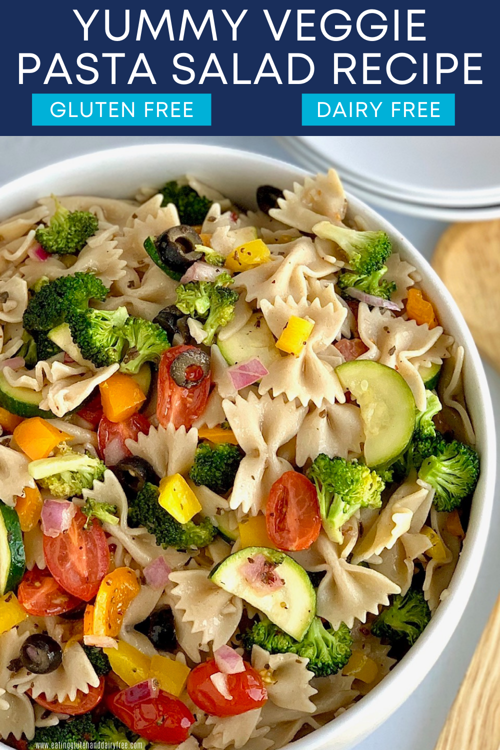 A large white serving bowl of bow tie pasta noodles, diced bell peppers, broccoli, olives, cucumbers, and purple onion with dressing.
