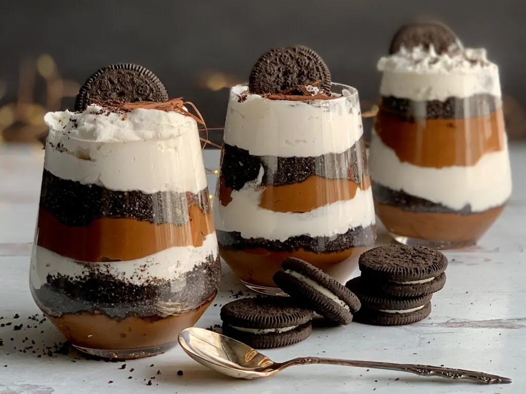 3 glass jars lined up diagonally layered with a thick homemade chocolate pudding, crushed oreo-like cookies, and a creamy white whipped topping. Then on the very top is more crush cookie, an oreo-like cookie, and chocolate curls. Next to the glass jars are more Oreo-like cookies.