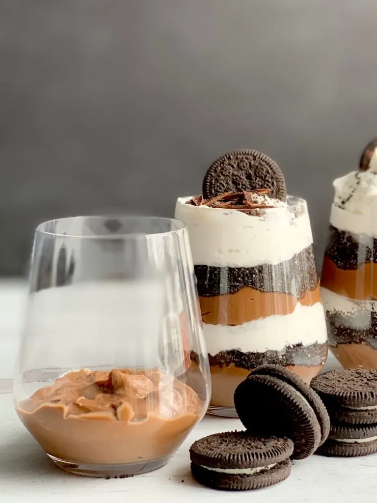 1 glass jar with the first layer of thick chocolate pudding on the bottom next to 2 glass jars layered with a thick homemade chocolate pudding, crushed oreo-like cookies, and a creamy white whipped topping. Then on the very top is more crush cookie, an oreo-like cookie, and chocolate curls. Next to the glass jars are more Oreo-like cookies.