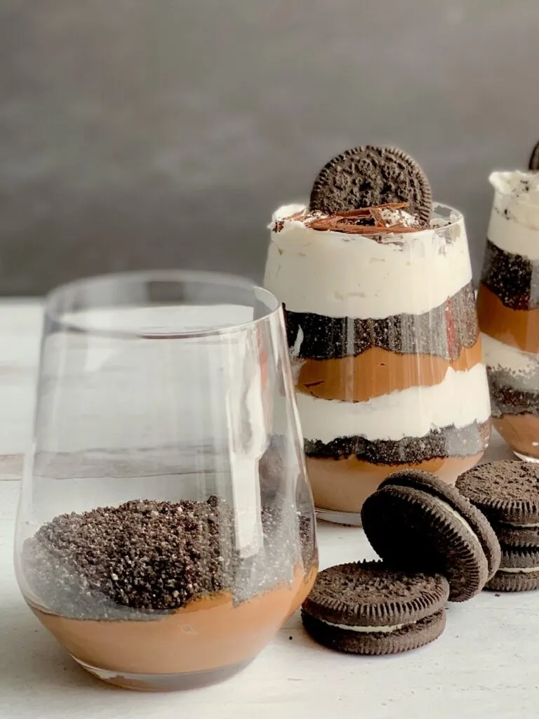 1 glass jar with the first layer of thick chocolate pudding on the bottom and cookie crumbs next to 2 glass jars layered with a thick homemade chocolate pudding, crushed oreo-like cookies, and a creamy white whipped topping. Then on the very top is more crush cookie, an oreo-like cookie, and chocolate curls. Next to the glass jars are more Oreo-like cookies.