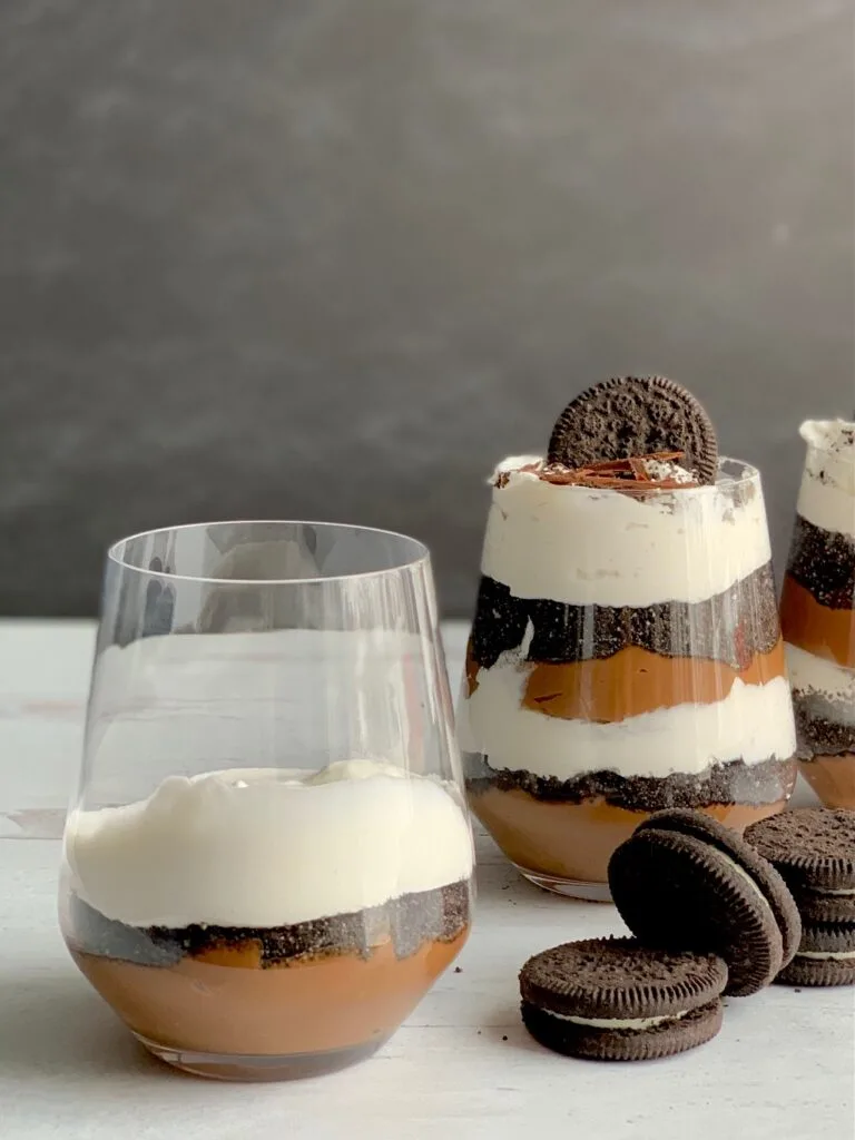 1 glass jar with the first layer of thick chocolate pudding on the bottom, then, cookie crumbs, and whipped topping next to 2 glass jars layered with a thick homemade chocolate pudding, crushed oreo-like cookies, and a creamy white whipped topping. Then on the very top is more crush cookie, an oreo-like cookie, and chocolate curls. Next to the glass jars are more Oreo-like cookies.