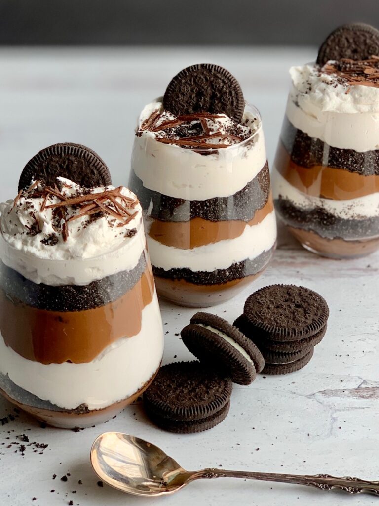 3 glass jars layered with a thick homemade chocolate pudding, crushed oreo-like cookies, and a creamy white whipped topping. Then on the very top is more crush cookie, an oreo-like cookie, and chocolate curls. Next to the glass jars are more Oreo-like cookies.