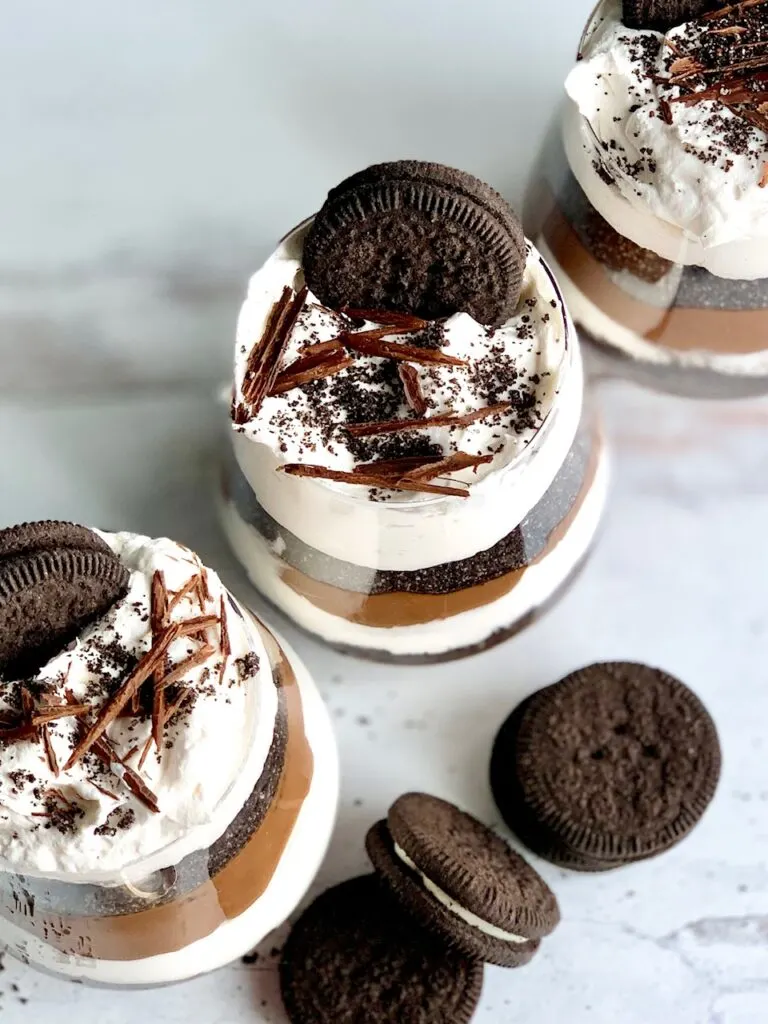 The top view looking down of 3 glass jars lined up diagonally layered with a thick homemade chocolate pudding, crushed oreo-like cookies, and a creamy white whipped topping. Then on the very top is more crush cookie, an oreo-like cookie, and chocolate curls. Next to the glass jars are more Oreo-like cookies.